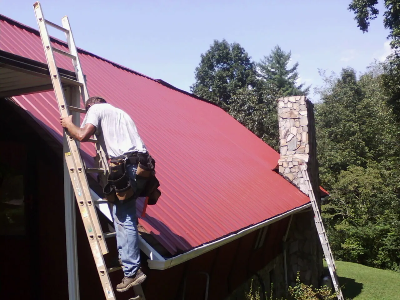 Roofer on a ladder beside a red metal roof and stone chimney — Soffit & Fascia Repair by Roofing Brothers in Reynolds
